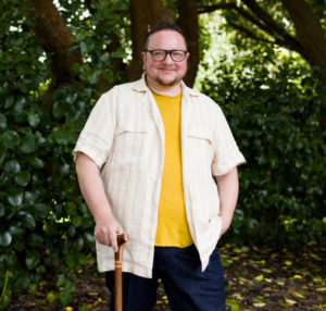 Image Description: A white, non-binary person, with short brown hair and facial hair, wearing blue jeans, a dark yellow t-shirt, a flax-colored linen short-sleeved button down short and rectangular olive-colored glasses, holding a brown wooden cane, stands in front of shadows and trees. He is smiling and looking directly into the camera.